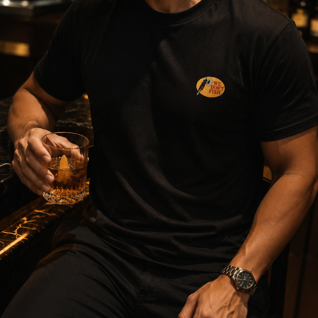 A man wearing a black short-sleeve t-shirt with a small circular gold and red We Dont Fish graphic on the left chest, sitting in a dimly lit bar environment and holding a glass of amber liquor.
