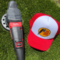 A cordless angle grinder with a red and black handle and a circular sanding disc attached, placed on green grass next to a red and white trucker hat featuring a fishing logo that reads 'We Dont Fish'.
