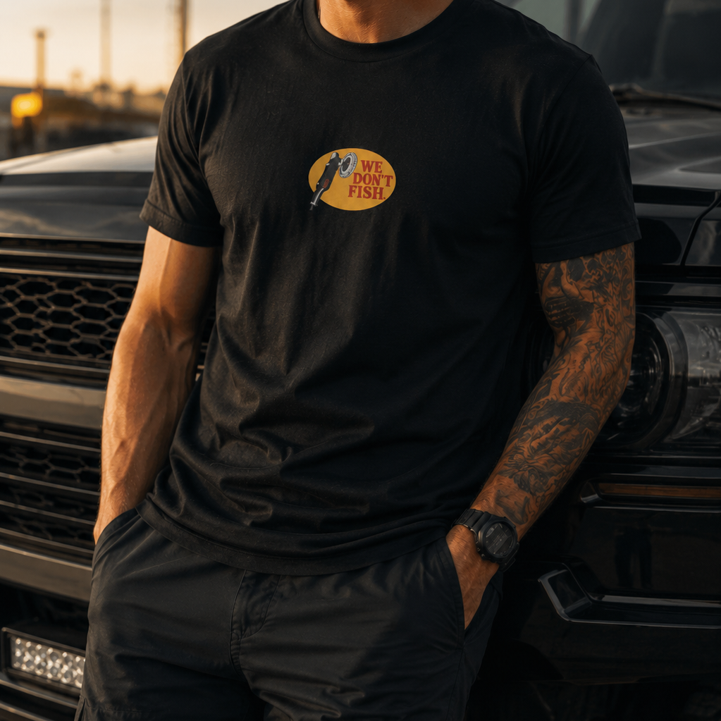 A man wearing a black crewneck t-shirt with a circular yellow and blue We Dont Fish graphic on the chest, standing next to a black truck at sunset.
